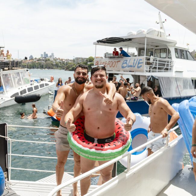 A group of people enjoying a sunny day on a boat party. The foreground shows two men, one wearing inflatable watermelon rings. Other boats and people swimming are visible in the background. The scene is lively and festive.