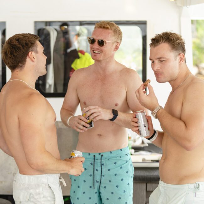 Three men in swim trunks enjoy snacks and beverages on a boat. One man wears sunglasses and smiles, while the others engage in conversation. They stand against a backdrop of water and trees, suggesting a relaxed, sunny atmosphere.