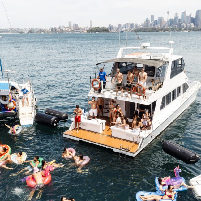 A party on boats with people enjoying in the water. Two yachts are anchored close together, with people on board and in the ocean on colorful inflatables. The city skyline is visible in the background under a clear sky.