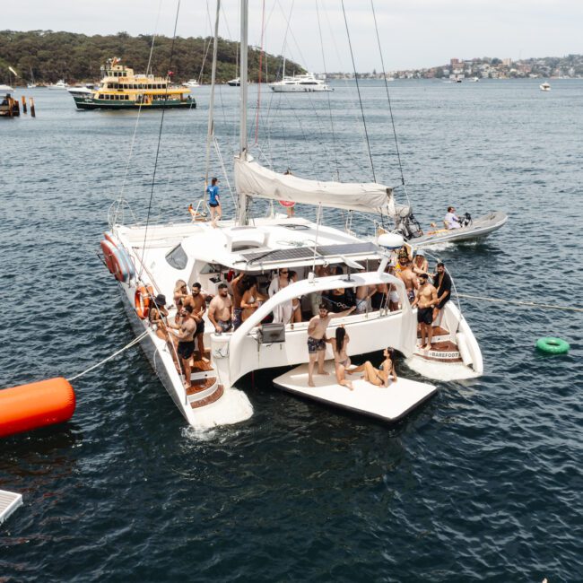 A group of people are enjoying a day on a large white catamaran boat anchored in calm waters. Some are sunbathing, while others stand and chat. Colorful buoys are floating nearby. In the background, there's a ferry and lush greenery on the shore.