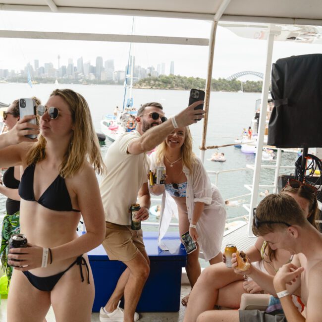 A lively group of people on a boat party, holding drinks and taking selfies. The scene includes a background view of a bay, dotted with other boats, and a city skyline visible in the distance. The atmosphere appears festive and summery.