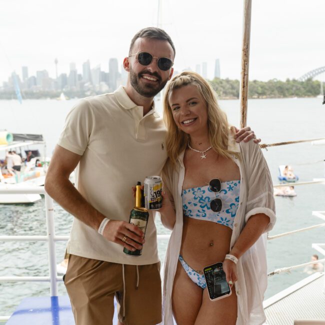 A man and woman smiling on a boat, both holding drinks. The man is wearing sunglasses and a light polo shirt, while the woman is in a blue-patterned bikini and cover-up, also holding a phone. Boats and a city skyline are visible in the background.