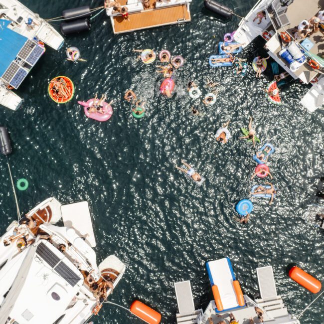 Aerial view of multiple boats gathered in water, with people swimming and using colorful inflatables. Some individuals are on the boats while others enjoy the water, creating a lively summertime scene.