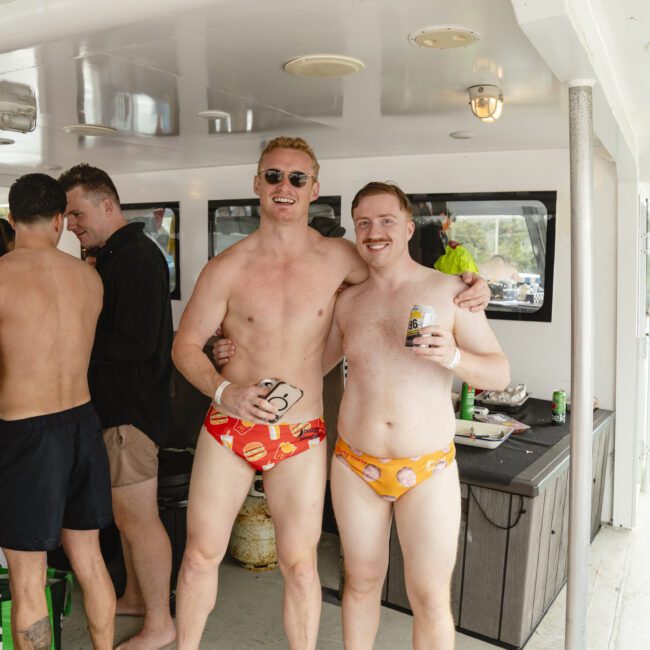 Two men wearing themed swim trunks pose together on a boat. One wears sunglasses, and both are smiling. Other people are on the boat in the background, including one in a black shirt and another in dark shorts.