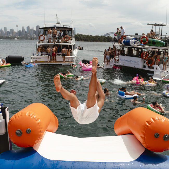 A person is mid-air, having jumped from an inflatable slide into the water. Several people are on boats and inflatables nearby, enjoying a sunny day on the water. The city skyline is visible in the background.