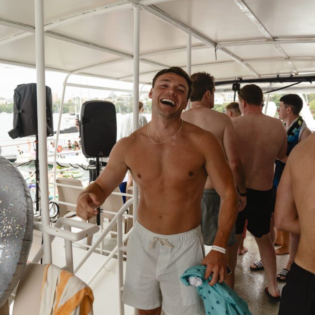 A shirtless man smiling and standing on a boat, surrounded by other people enjoying a sunny day. He is holding a towel and wearing swim trunks. Other passengers are socializing in the background, with water and boats visible outside.