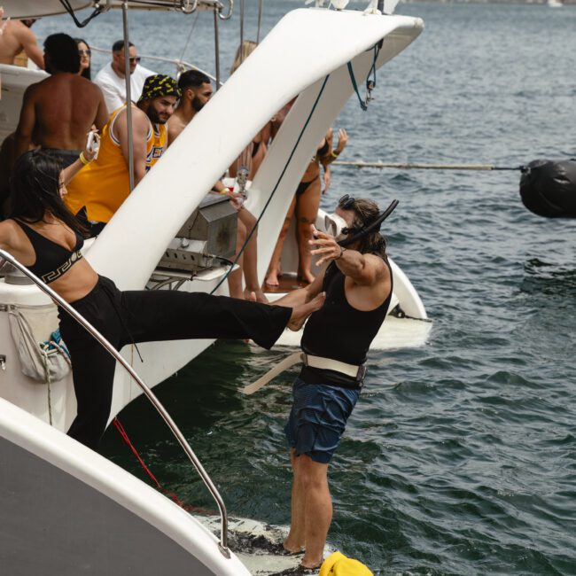 A man wearing swim trunks playfully pretends to fall backward from a boat while holding the foot of a woman standing on the deck. Several people in swimwear are nearby, and a person in a yellow cap swims in the water.