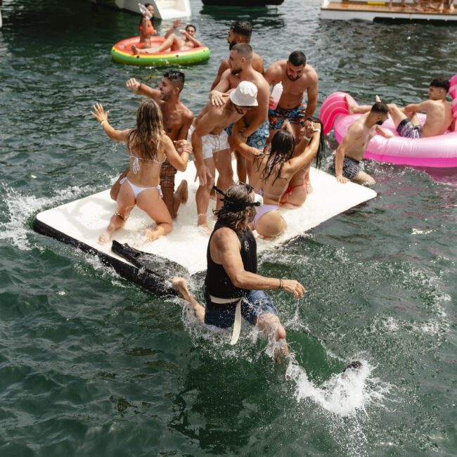 People enjoy a lively boat party on a lake, with some dancing on a floating platform and others swimming nearby. The background features more people on boats and colorful inflatables. The scene is vibrant and filled with summer fun.