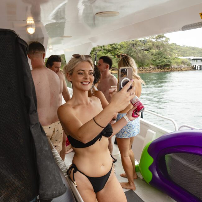 A woman in a black bikini takes a selfie on a boat with a group of people in swimwear around her. The boat appears to be docked near a lush, green shoreline. Inflatable items are visible on the deck.