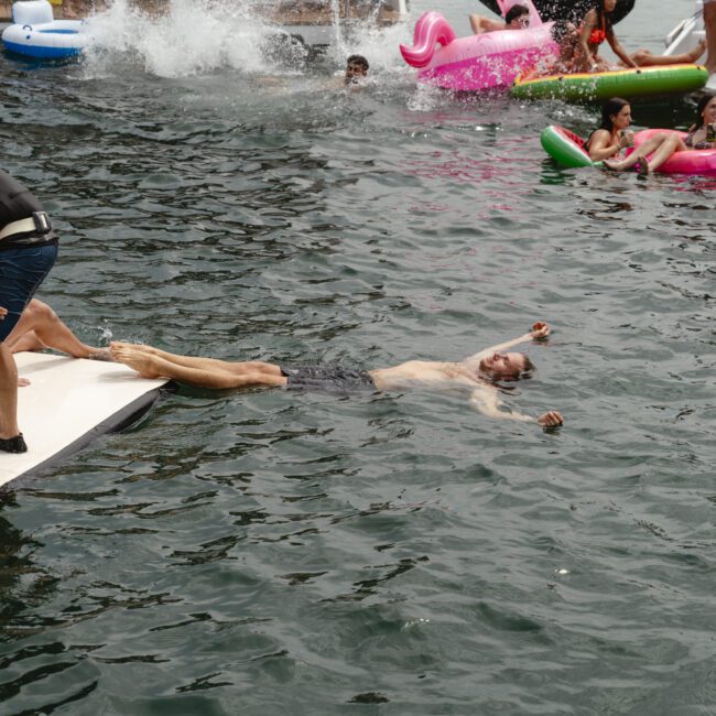 A group of people enjoying a day on the water with colorful inflatables. One person floats on their back in the water, while others lounge nearby on a platform. Boat visible in the background.