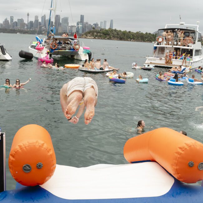 A person is mid-dive from an inflatable platform into the water, surrounded by people swimming and floating on inflatables. Multiple boats and a city skyline are visible in the background under a cloudy sky.