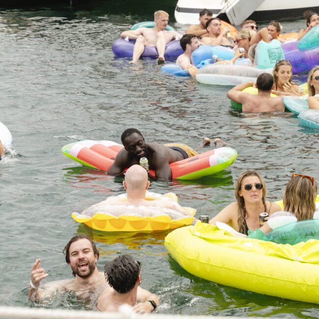 A lively group of people relax on colorful inflatables in a body of water, enjoying a sunny day. Some are lying down, others are sitting in the floats, surrounded by more bathers and boats in the background.