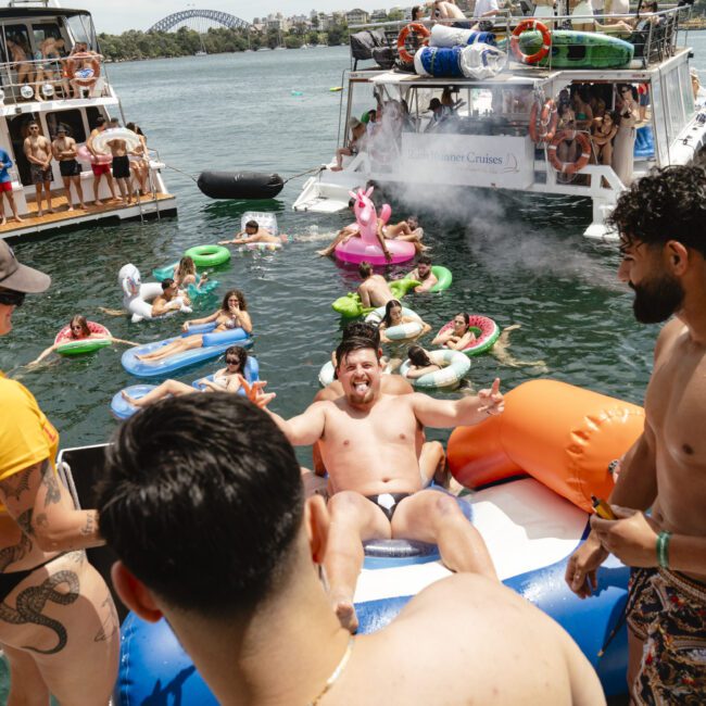 A lively boat party on the water with people enjoying music, drinks, and floating on inflatable rafts. The background features a large boat and a bridge against a clear sky.