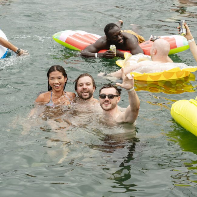 A group of people enjoying a day in the water. Some are on inflatable floats, while others are swimming and smiling for the camera, holding drinks. The setting is lively and relaxed.