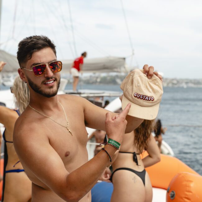 A man wearing sunglasses is smiling and pointing at a beige cap with the name "Gustavo" on it while on a boat. He is shirtless and surrounded by other people wearing swimsuits, with a body of water and another boat in the background.