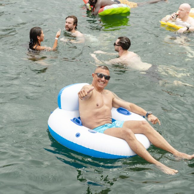 A group of people enjoying a sunny day in a lake. A man in sunglasses is smiling and lounging on a blue and white inflatable chair, while others swim and chat around him. The water is calm and refreshing.