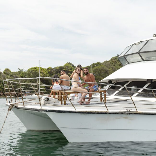 A group of people enjoying a sunny day on the deck of a white boat. They are surrounded by calm water and lush greenery in the background. The sky is overcast, and the atmosphere appears relaxed and cheerful.