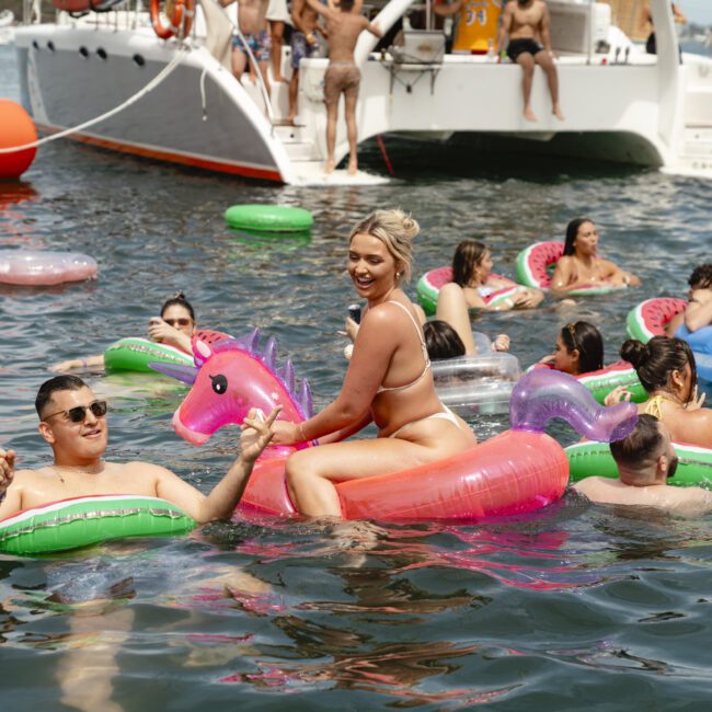 A group of people enjoying a sunny day on the water with inflatable pool floats. They are smiling and laughing, surrounded by boats. One person is sitting on a pink unicorn float, while others relax on various colorful inflatables.