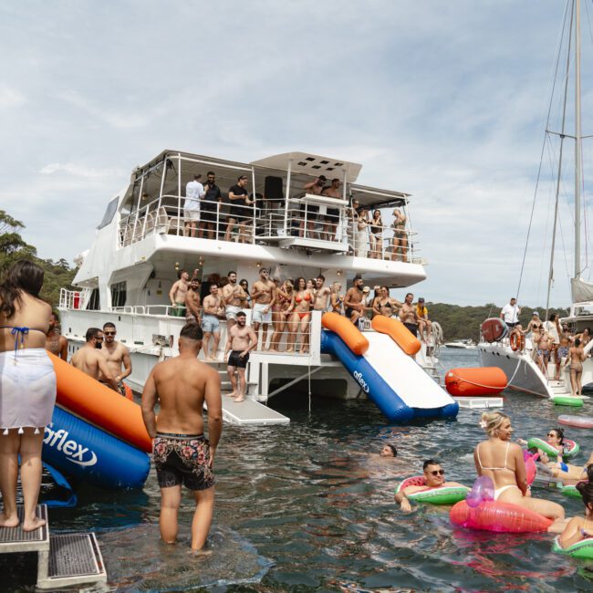 A large group of people enjoy a boat party in sunny weather. Some are on the yacht with inflatable slides, while others relax on colorful floaties in the water. Another boat is anchored nearby. The scene is lively and festive.