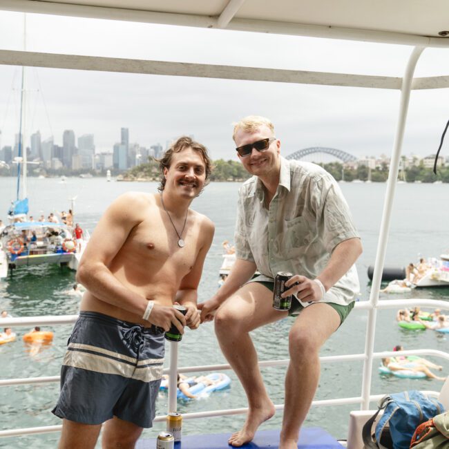 Two men are on a boat during a festive gathering on the water. Both are casually dressed, holding drinks, and smiling at the camera. In the background, other boats and people enjoying the event can be seen with a city skyline under a cloudy sky.