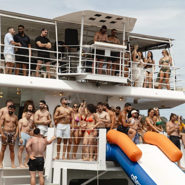 A group of people in swimwear are gathered on a multi-tiered boat with a water slide. Some are standing, others are seated, and some are dancing. The boat is on a body of water, surrounded by green trees in the background.