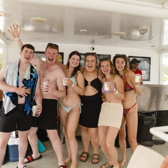 A group of six people in swimwear pose and smile on a boat, holding drinks. Others are visible in the background, also in swimwear, enjoying a day on the water.