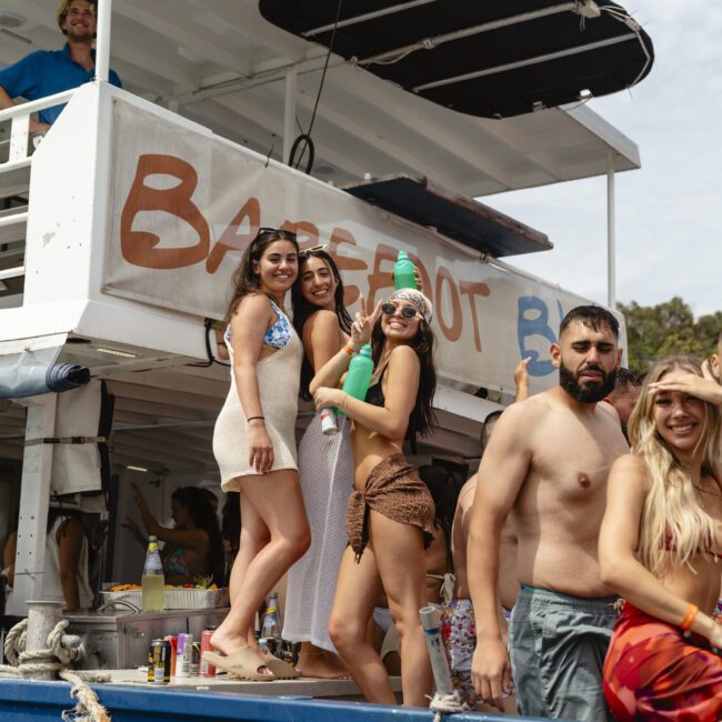 A group of people enjoying a boat party. Three women are posing together, with one holding a green inflatable toy. A man and another woman stand nearby, all smiling. The boat is labeled "Barefoot." They appear to be having a good time.