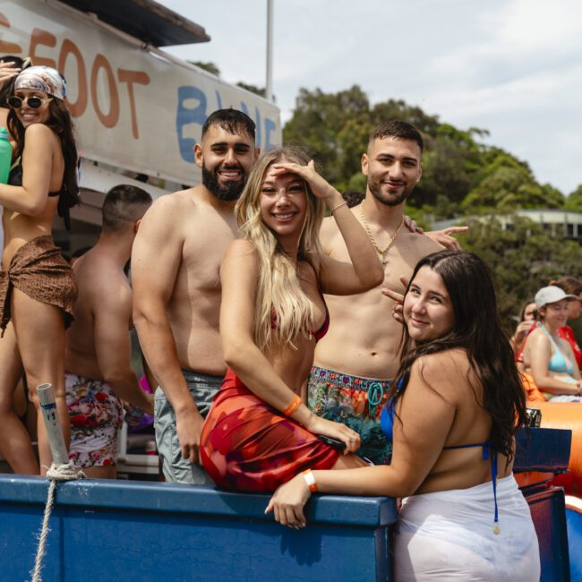 A group of four people in swimwear pose on a boat. Two are shirtless men, and two are women in bikinis and skirts. They appear to be enjoying a sunny day, with trees and water in the background.