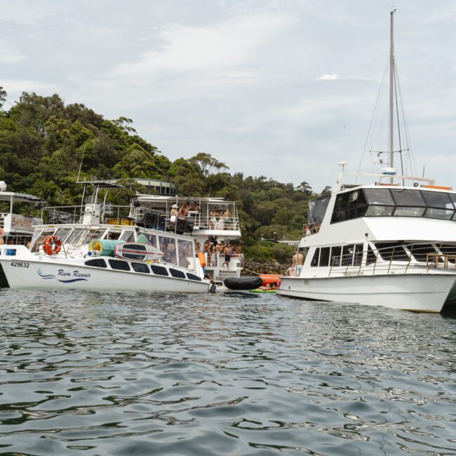 Several boats are moored together on a calm body of water, surrounded by lush green hills. People are visible aboard the boats, enjoying the scenery. The sky is overcast, creating a serene atmosphere.