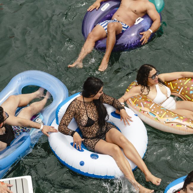 Four people are lounging on inflatable pool tubes in the water. They are relaxing and enjoying a sunny day, with the floats in various shapes and colors, including a donut and an ice cream design.