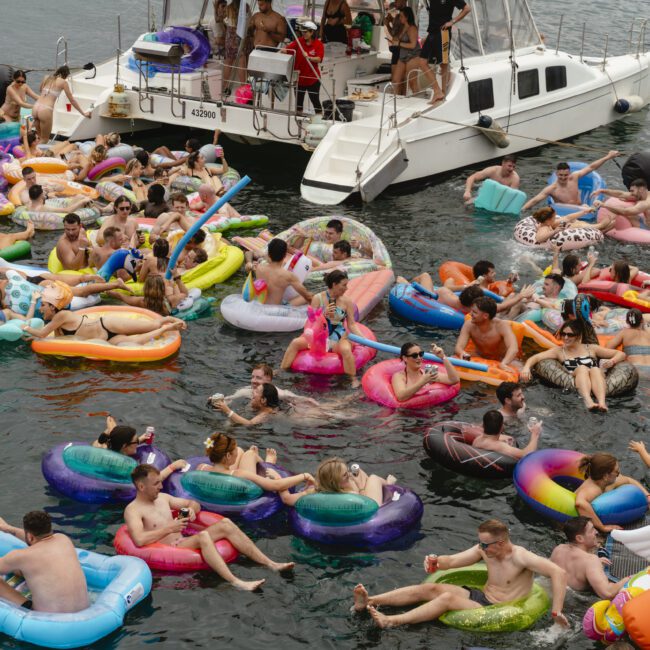 A lively scene of people enjoying a party on colorful inflatable floaties in the water near a boat. They are laughing, talking, and relaxing in the sun, creating a festive atmosphere with a mix of vibrant pool toys and swimwear.