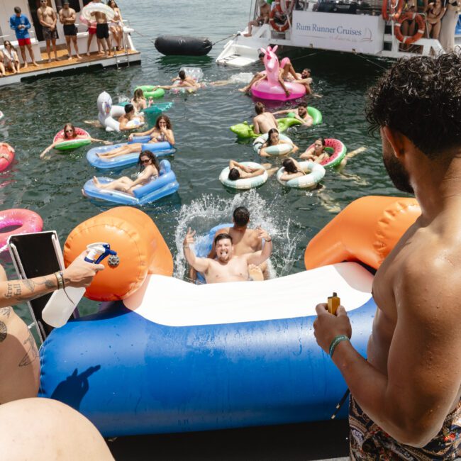 A lively pool party on a boat features people enjoying various colorful inflatables in the water. Some are sliding into the water, while others relax on floating toys. A few people stand on the deck, observing the fun. "Sail Gustavo" is visible in the corner.