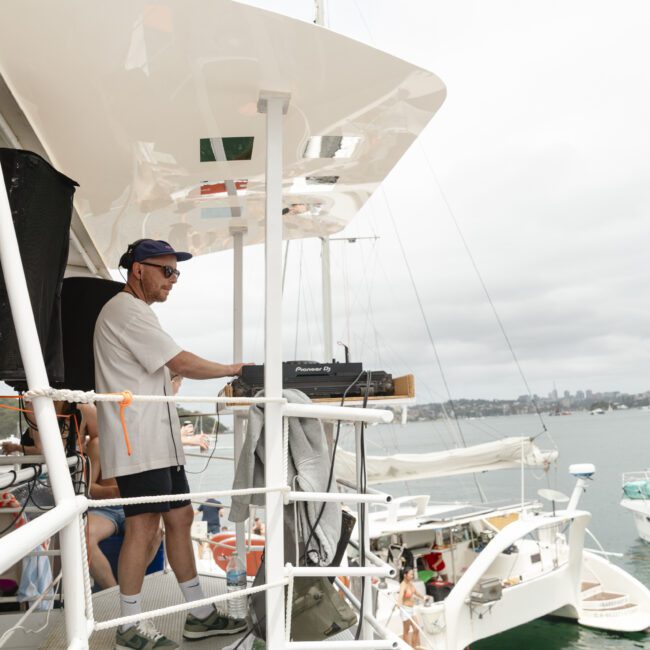 A person in shorts, a T-shirt, cap, and sunglasses is DJing on a boat deck, surrounded by lake scenery and other boats. The sky is cloudy, and people are visible in the water below.