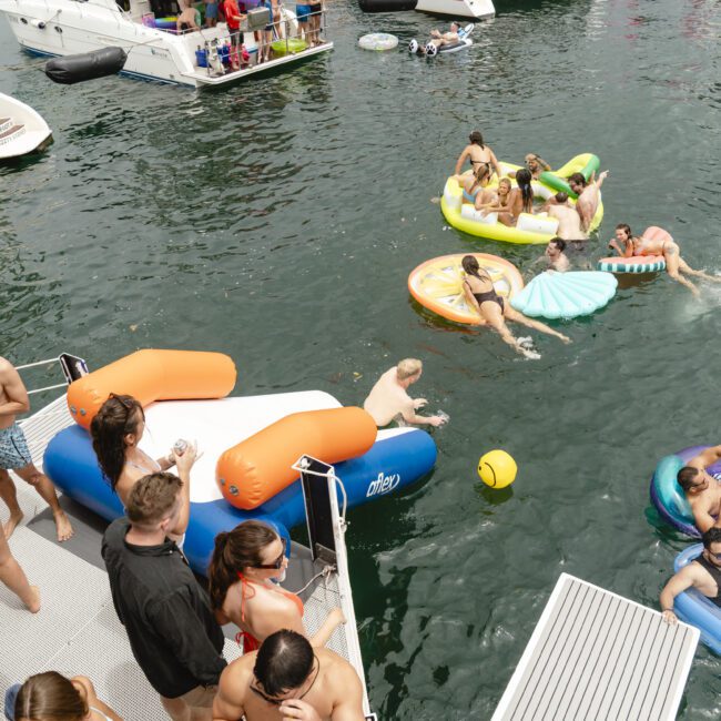 A lively scene of people enjoying a sunny day on a lake with several boats. Individuals are swimming, lounging on inflatable floats, and socializing near the water. The atmosphere is relaxed and festive.