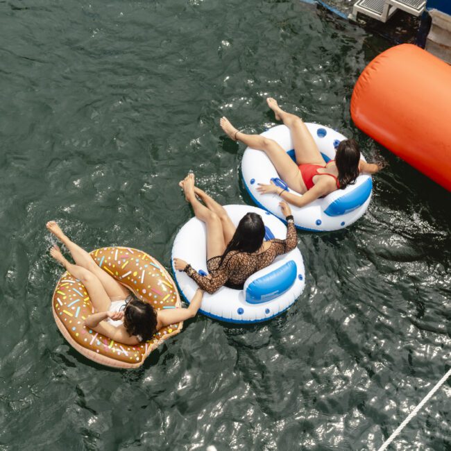 Three people relax on inflatable rings in the water, holding hands. One floats on a donut-shaped ring, while the others use round floaties. A large orange buoy is nearby, with part of a boat and dock visible.