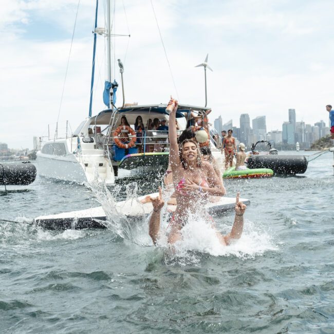 A group of people enjoying a day on the water with a boat in the background. In the foreground, two people are energetically playing in the water. The city skyline and a partly cloudy sky are visible in the distance.