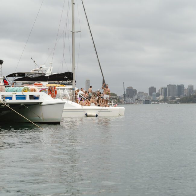 Boats anchored on a calm body of water with people relaxing on board. The skyline of a city with skyscrapers is visible in the background under a cloudy sky.