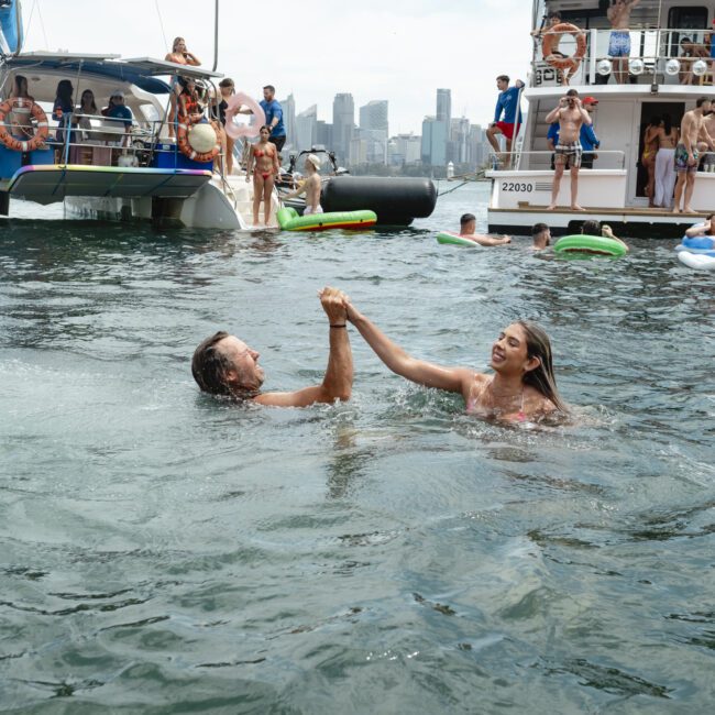 Two people holding hands while swimming in a body of water, surrounded by boats and people on inflatable floats, with a city skyline in the background.