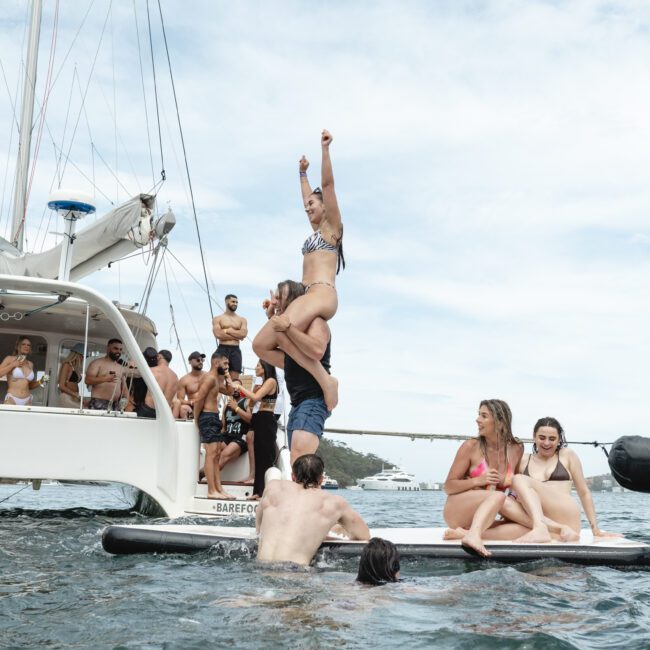 A group of people enjoying a day on the water. Some are on a boat, while others are on a paddleboard. One person stands triumphantly on another's shoulders. They are all dressed in swimwear and appear to be having fun.