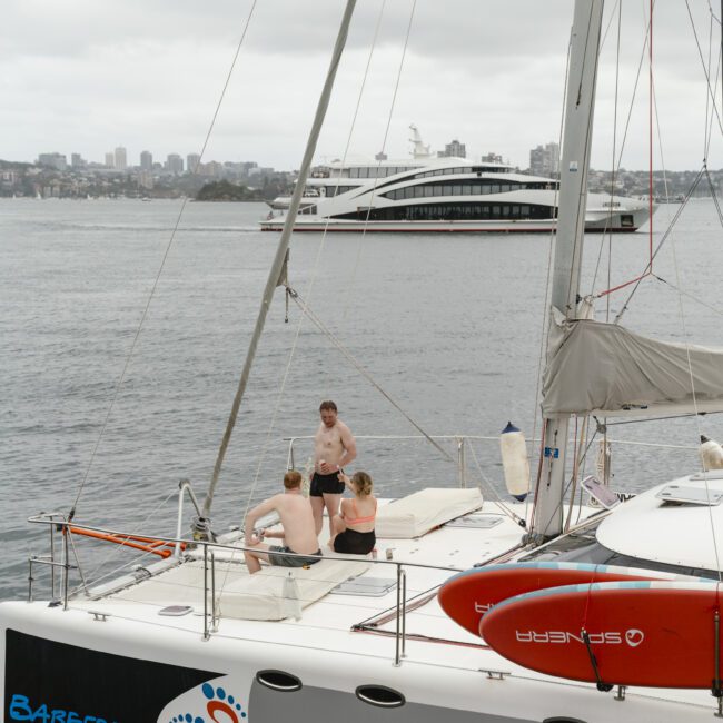 Three people sit and relax on the deck of a sailboat named Barefoot, anchored in calm waters. The city skyline and another large boat are visible in the background under a cloudy sky.