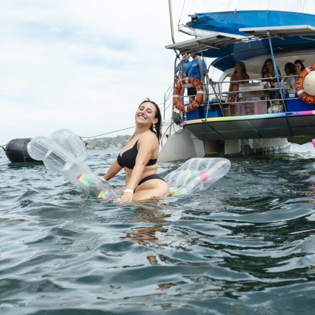 A person in a black swimsuit is smiling and sitting on an inflatable float in the water, with a boat and people in the background. The sky is cloudy.