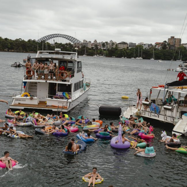 A lively scene with people floating on colorful inflatables in a harbor, near two anchored boats. The background features a bridge and cityscape under a cloudy sky.