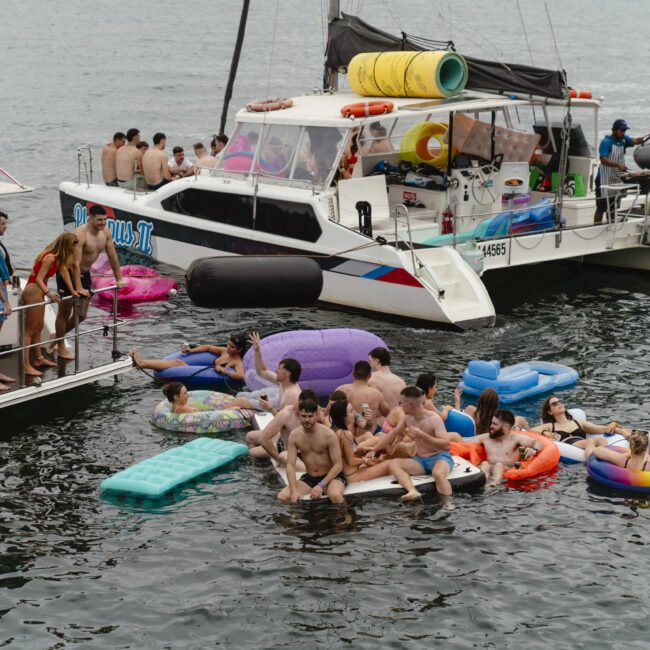 People enjoying a party on a boat and in the water, surrounded by colorful inflatables including a flamingo and a unicorn. The scene is lively, with groups socializing on the boat and in the water, creating a festive atmosphere.