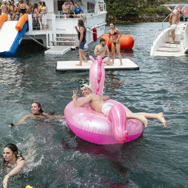 A man lounges on a pink flamingo float, surrounded by people swimming and playing in the water. A boat and inflatable slides are in the background, with more people enjoying the scene. The atmosphere is lively and festive.