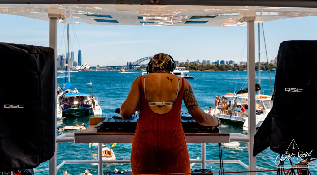 A DJ wearing headphones performs on a boat with a scenic backdrop of a city skyline and a bridge. Several boats and swimmers are visible on the blue water under a clear sky.
