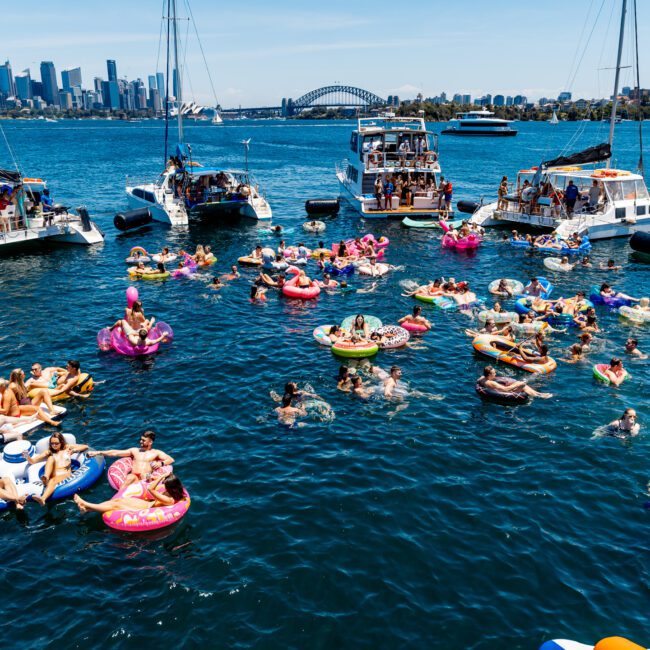 A vibrant scene of people on colorful inflatable floats in a bay, surrounded by yachts. The city skyline is visible in the background under a clear blue sky.