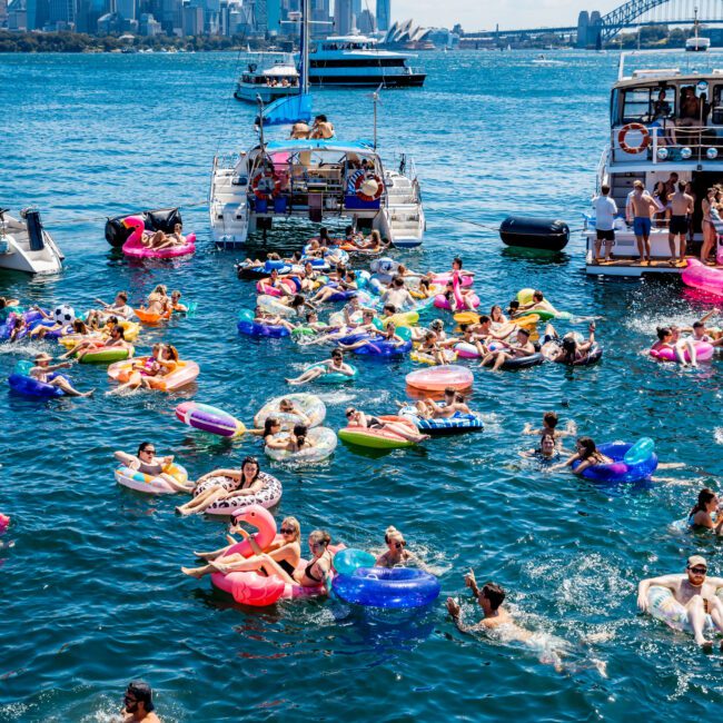 People enjoying a sunny day floating on colorful inflatables in a bay, with boats anchored nearby. The city skyline and a distinctive bridge are visible in the background.