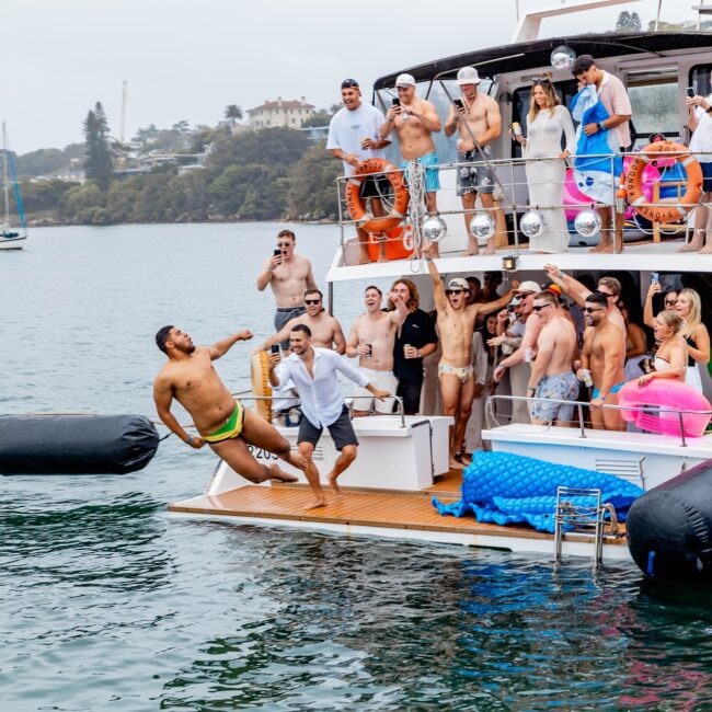 A lively social club gathers on the deck of a docked yacht, where laughter fills the air. One adventurous soul leaps into the calm water as others cheer them on. The shoreline's trees and houses provide a picturesque backdrop to this festive celebration.