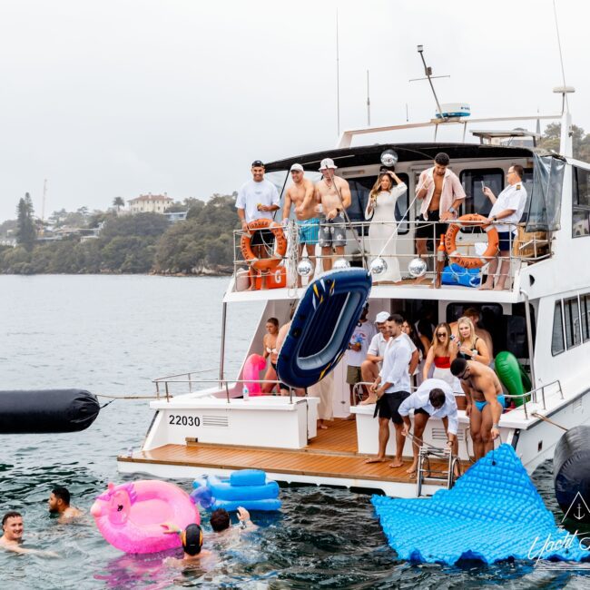 A group of people from the social club are enjoying a day on the white yacht, with several standing on the deck and others swimming and floating on colorful inflatables in the water. The background shows a calm body of water and distant trees.