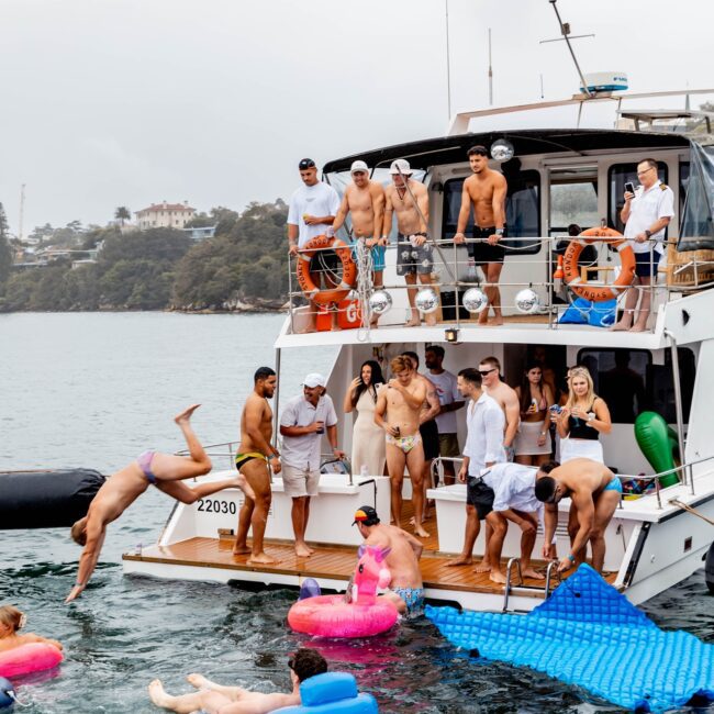 A lively social club enjoys a day on the yacht; some members leap into the water while others relax on inflatable floats. The yacht is anchored near a scenic, wooded shoreline under an overcast sky.